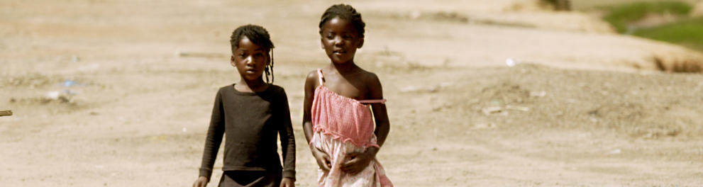 Duas crianças negras andam, descalças, em um chão de terra.