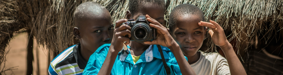 Três crianças africanas estão de frente para quem bateu a foto. A criança do meio está com uma câmera fotográfica no rosto, como se estivesse batendo uma foto também