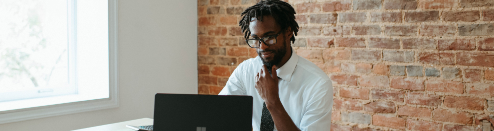 Um homem negro de camisa social e gravata está sentado e olhando para um notebook aberto em sua frente