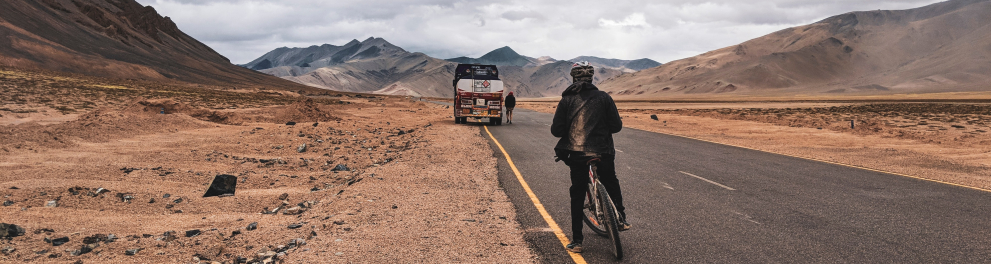 Um ciclista está em pé ao lado de sua bike em uma estrada muito comprida e rodeada por rochas e solo arenoso. Mais à frente na estrada há um furgão ou caminhão estacionado com uma pessoa ao lado