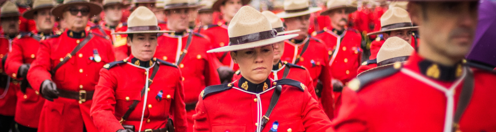 Vários soldados canadenses estão enfileirados e marchando. Eles estão usando a típica roupa vermelha canadense, luvas e calças pretas e o chapéu bege