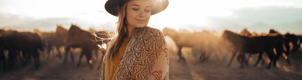 Uma mulher está de olhos fechados e de frente para quem bateu a foto. Ela está trajando uma roupa country, com chapéu de cowgirl e há vários cavalos ao fundo da imagem