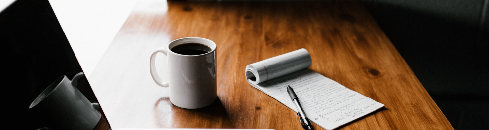 Um notebook aberto e desligado, uma xícara branca de café, um bloco de anotações, uma caneta e um celular estão em cima de uma mesa de madeira sendo iluminada pela luz solar que vem da janela ao lado