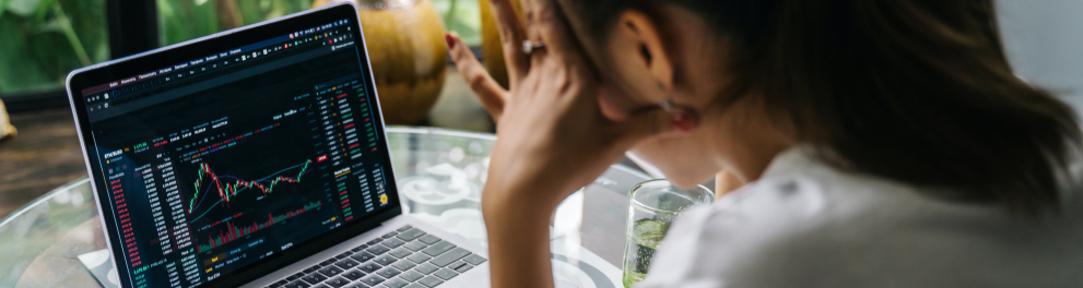 Uma mulher de costas para quem bateu a foto está com a mão esquerda na cabeça como se estivesse preocupada com algo enquanto olha o notebook ligado em uma tela de gráficos econômicos