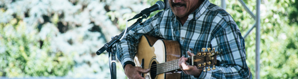 Um homem de bigode e chapéu de cowboy está sentado tocando violão e cantando algo com um microfone à sua frente
