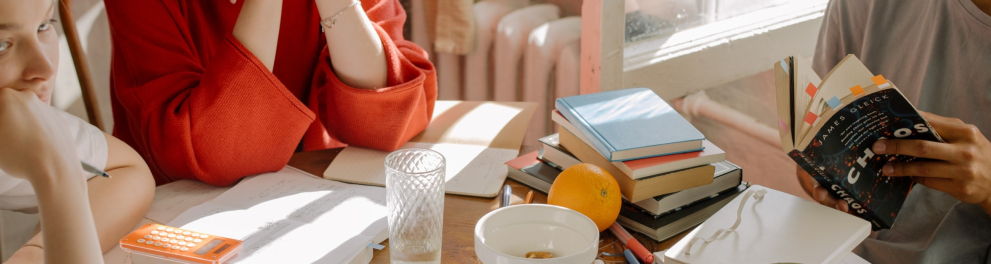 A imagem mostra três pessoas sentadas em uma mesa com vários livros, cadernos e papeis