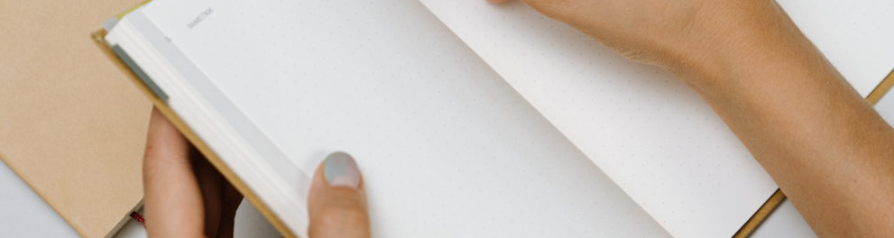 Imagem das mãos de uma pessoa e um caderno. A pessoa está segurando o caderno com a mão esquerda e prestes a escrever algo com uma caneta segurada pela mão direita