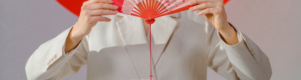 Uma mulher oriental está segurando um leque com as duas mão, à frente de seu rosto, abaixo da altura dos olhos, de frente para quem bateu a foto. Atrás dela está a bandeira do Japão