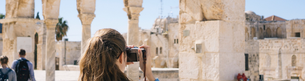 Uma mulher está de costas para quem bateu a foto e tirando uma fotografia de ruínas que parecem ser em algum local de Israel