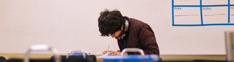 A imagem mostra um estudante em uma sala de aula concentrado em um papel