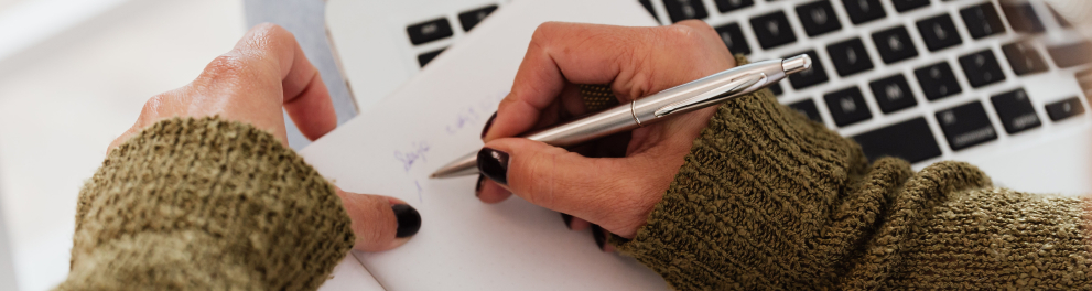 Zoom nas mãos de uma pessoas branca que está escrevendo algo em um bloquinho de papel e há um notebook ligado ao lado do bloco