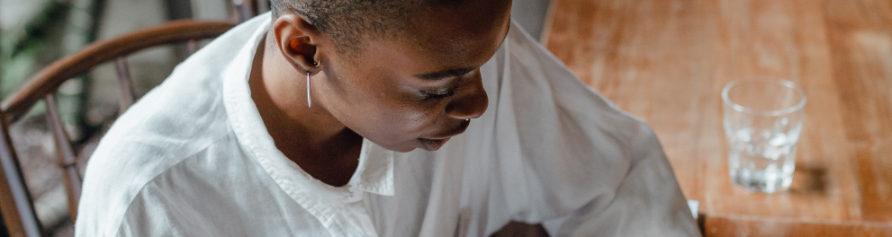 Uma mulher negra está sentada escrevendo em uma folha de pape apoiada em uma mesa de madeira, que também tem um celular em cimal