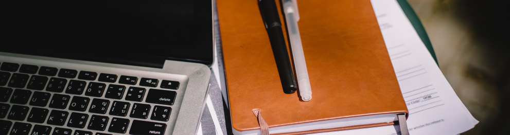 Duas canetas estão em cima de um caderno, que está ao lado de um notebook aberto, porém desligado