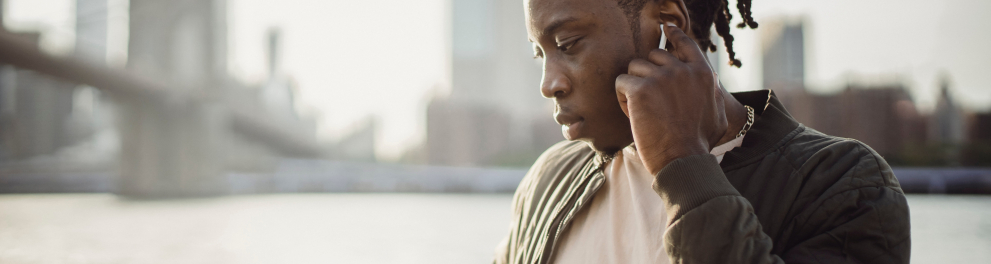 Um homem está ao ar livre com um celular na mão direita e fone de ouvido bluetooth na orelha esquerda. Ele está com o dedo indicador da mãok esquerda tocando neste fone. Ao fundo, podemos notar vários prédios e é claramente a cidade de Nova Iorque