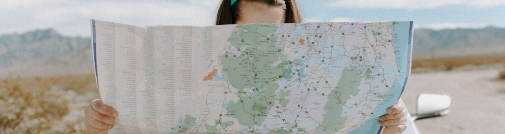 Uma mulher branca está segurando um mapa de papel com as duas mãos e olhando para ele. Ela está em pé, encostada ao capô de um carro em meio à uma estrada bem deserta de dia