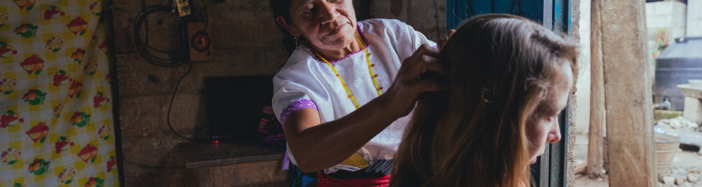 Uma mulher de mais idade com traços físicos de uma típica latina está mexendo nos cabelos de uma outra mulher mais jovem