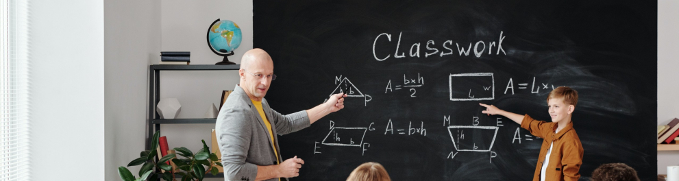 A imagem mostra uma sala de aula com um professor e um estudante em pé em frente a uma lousa