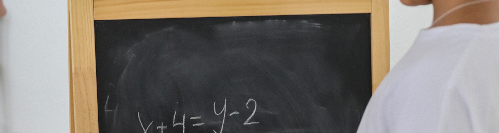 Uma menina está de costas para quem bateu a foto e de frente para um pequeno quadro negro com uma equação matemática para resolver