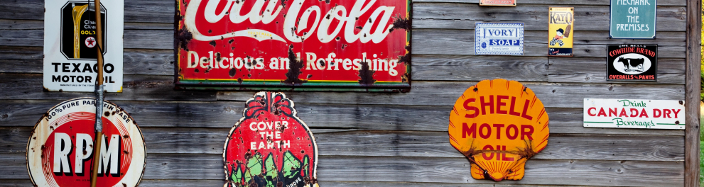 Cartazes de publicidade de marcas como Texaco, Coca-Cola, Shel, entre outras, colados em uma parede de madeira