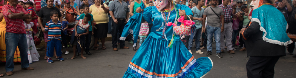 Duas pessoas com fantasias coloridas e máscaras nos rostos estão dançando na rua enquanto dezenas de outras estão olhando atentamente. É uma típica dança de algum país latino
