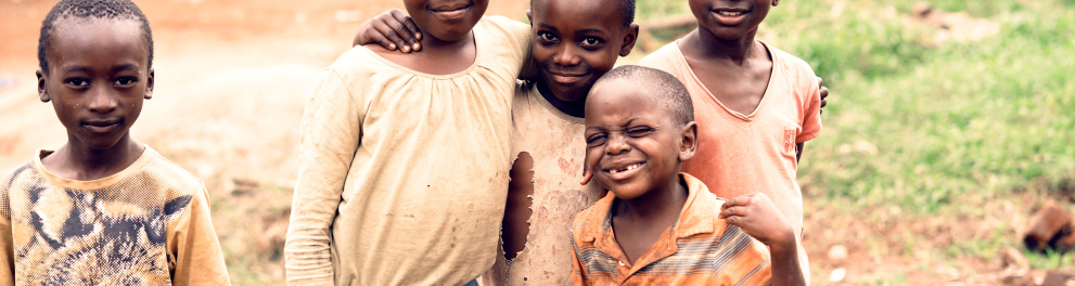 Cinco crianças africanas estão com roupas humildes (sujas e até rasgadas), abraçadas e olhando sorridentes para quem bateu a foto