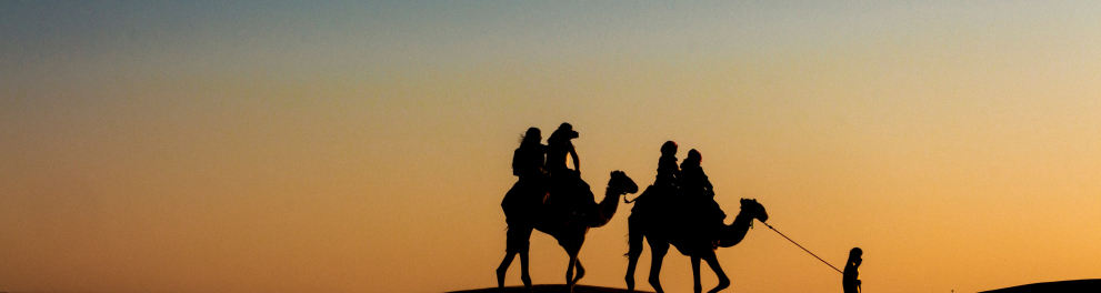 Imagem distante de dois camelos andando em um deserto com pessoas em suas corcovas 