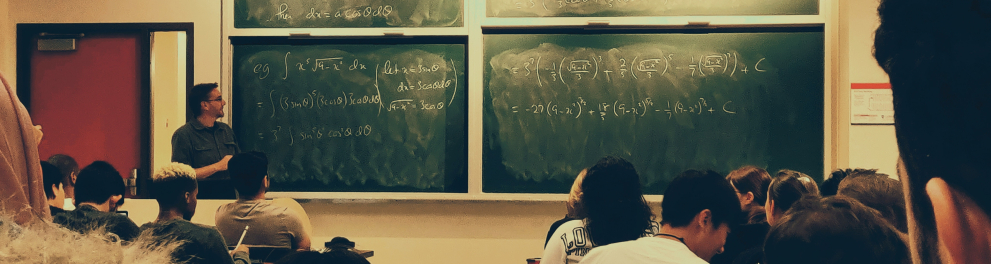 Uma sala de aula com os alunos sentados de costas para quem bateu a foto e o professor lá ao fundo da imagem, em frente ao quadro negro, falando algo