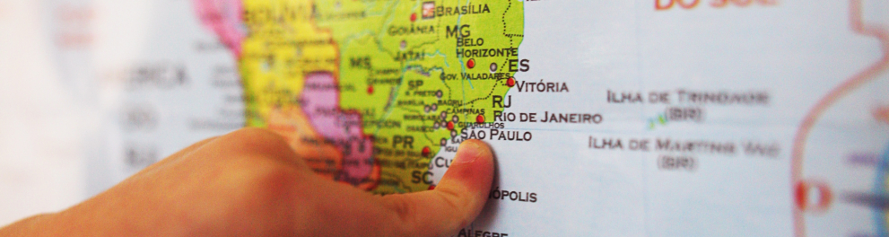 A mão esquerda de uma pessoa branca está tocando com o dedo indicador em um mapa da América do Sul, mais precisamente no mapa brasileiro