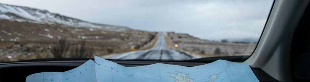 Imagem feita do interior de um carro, mais precisamente do banco carona. Mostra um mapa de papel apoiado no painel do carro e a estrada à frente através da janela frontal do veículo