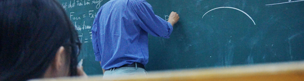 Um professor está de costas para quem bateu a foto e está escrevendo no quadro negro, onde já há algumas frações escritas 