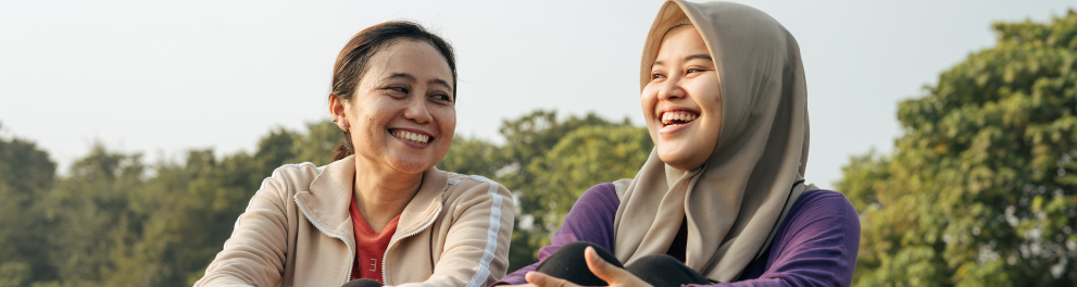Duas mulheres asiáticas estão sentadas em um campo aberto com muitas árvores ao fundo. Elas estão sorridentes e parecem estar conversando.