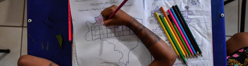 Uma criança está sentada em sua cadeira na sala de aula e está escrevendo em um caderno. A imagem é feita por cima da criança
