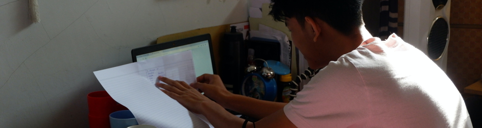 Imagem de um menino de costas para quem bateu a foto. Ele está sentado diante de um computador, livros e folhas e está claramente estudando