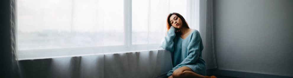 Uma jovem mulher está triste e sentada no chão de casa se apoiando na janela