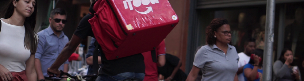 Imagem de um meio urbano, com várias pessoas andando em uma rua e um entregador está pedalando em sua bicicleta, com uma mochila de entrega do Ifood às costas