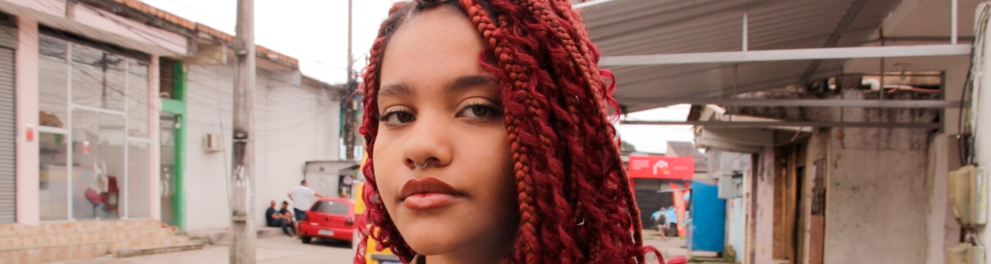 Uma jovem negra, com cabelo trançado em tom avermelhado, olha para quem a fotografa. Ao fundo, ruas e veículos em um dia cinzento.