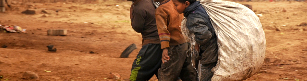 Três meninos estão vestidos com roupas humildes e sujas e carregando sacos pesados nas costas em meio a um solo arenoso e seco