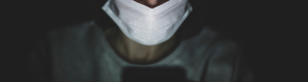 Um homem de máscara de prevenção contra a Covid está no escuro e com o celular ligado à sua frente....a única iluminação da foto é a luminosidade do aparelho celular