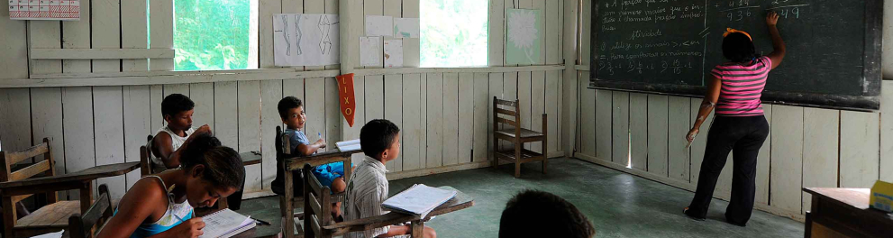 Uma sala de aula humilde, precária de infraestrutura e com poucos alunos sentados em suas cadeiras, enquanto a professora está escrevendo algo no quadro negro