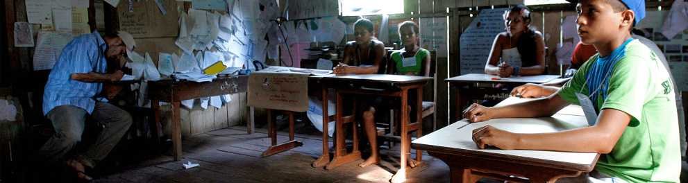 Uma sala de aula humilde, precária de infraestrutura e com poucos alunos sentados em suas cadeiras, enquanto a professora está escrevendo algo no quadro negro
