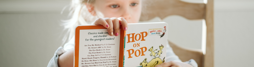 Uma jovem menina loira está sentada lendo um livro infantil, de frente para quem bateu a foto