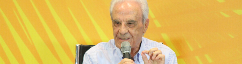 Miguel Arroyo, um senhor grisalho, está sentado e falando em um microfone