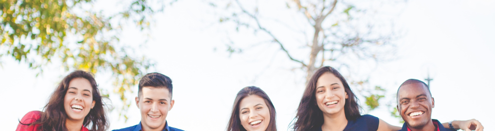 Cinco pessoas jovens estão sentadas lado a lado ao ar livre e sorridentes posando para a foto. São três mulheres e dois homens