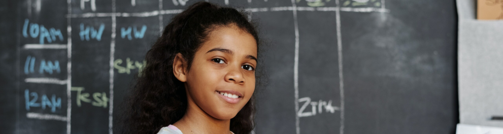 Uma menina sorri enquanto segura um caderno. Ao fundo, o quadro da sala de aula.