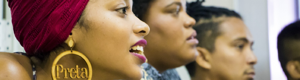 Foto pegando o perfil de três pessoas enfileiradas. A pessoa mas próxima da imagem é uma mulher negra e jovem com um brinco dourado que tem a palavra "preta" escrita ao centro