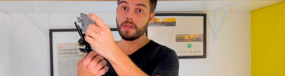 O professor Ney Mello, um homem branco, de barba e cabelos curtos está segurando um tripé e um celular enquanto fala algo