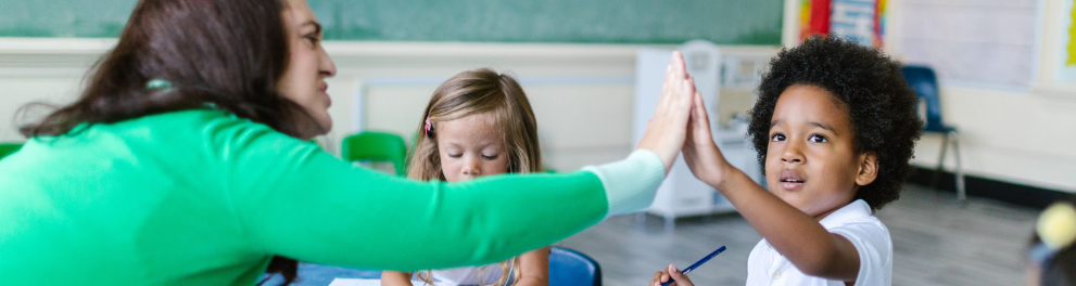 Três pessoas estão sentadas em uma sala de aula. À esquerda, uma mulher adulta saúda uma das outras duas crianças encostando a palma de sua mão na palma da mão da criança mais à direita.