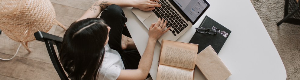 Uma mulher está sentada diante de um laptop, com alguns livros ao seu lado. A foto foi feita por cima dela.
