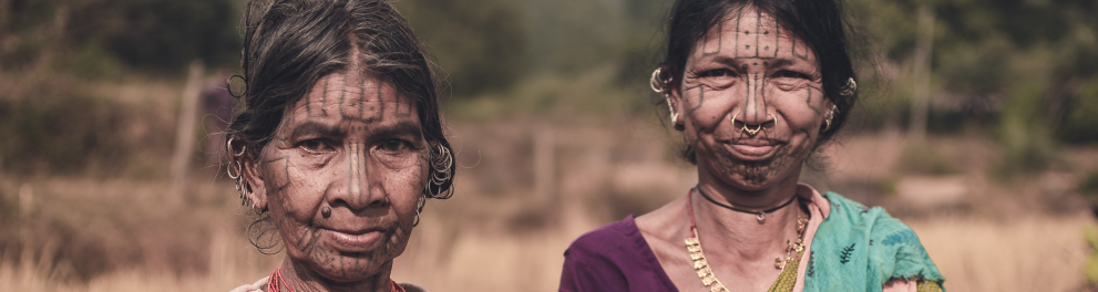 Duas mulheres indígenas olham para quem as fotografa. Ao fundo, um campo com mato ligeiramente seco.