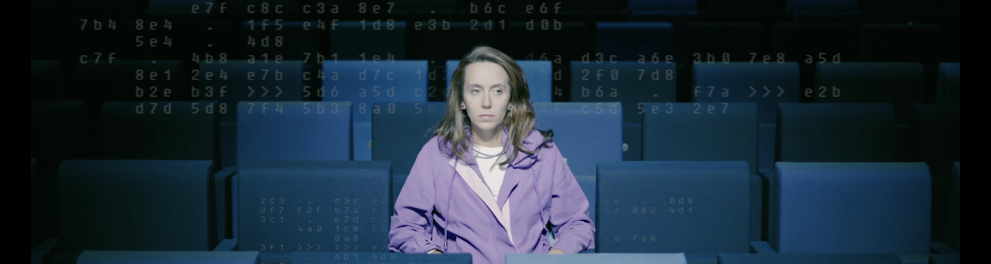Uma mulher está sozinha, sentada em uma sala de cinema escura, As poltronas são em tom azulado.